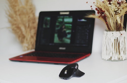 Mouse Logitech preto em mesa branca ao lado de notebook vermelho e jarro com plantas.