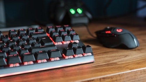 Mouse gamer preto com leds vermelhas ao lado de teclado gamer com leds vermelhas em cima de mesa de madeira.