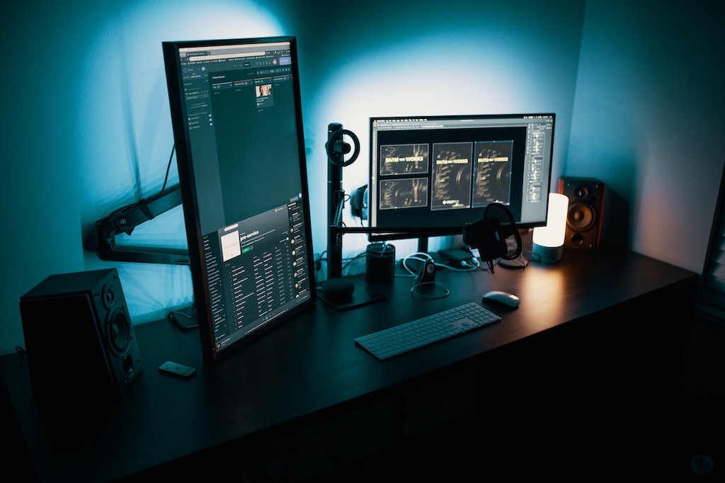 Computador com dois monitores, um na horizontal e um na vertical, em cima de uma mesa escura em um quarto com pouca iluminação.