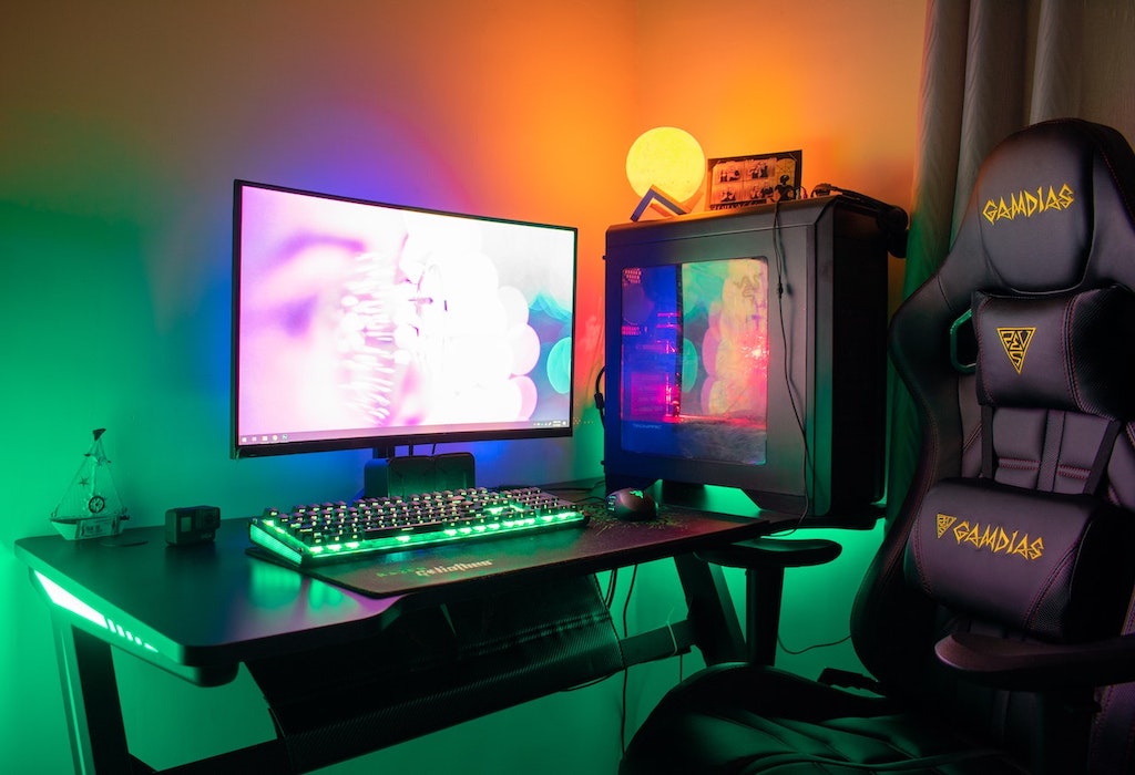 Setup gamer. Cadeira gamer preta e amarela ao lado de uma mesa com um computador gamer. Monitor com bastante brilho, teclado iluminado com led verde e gabinete transparente mostrando componentes iluminadas em led.