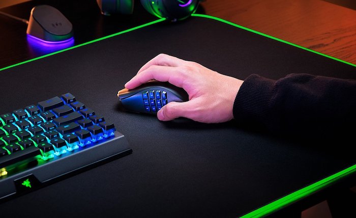 Homem usando um Mouse Razer preto com botões programáveis em um fundo preto ao lado de um teclado com leds.
