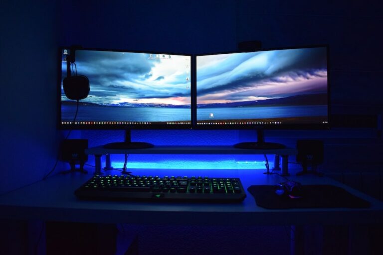 PC gamer com dois monitores numa base iluminada, teclado com led, headset pendurado no monitor em um quarto escuro.