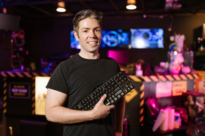 Homem de pele branca segurando um teclado mecânico preto, sorrindo, em um estúdio de jogos.