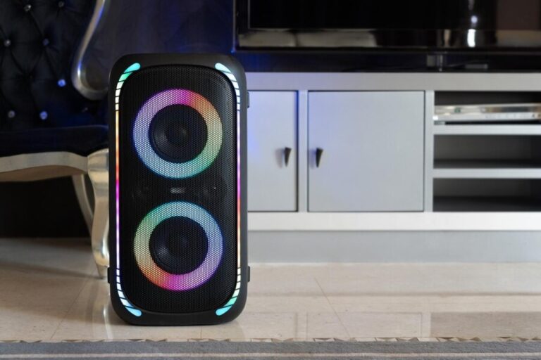 Caixa de som amplificada preta com dois alto-falantes e luzes LED coloridas, dentro de casa, em frente a uma poltrona azul de madeira. Atrás, vemos um armário branco de cozinha.